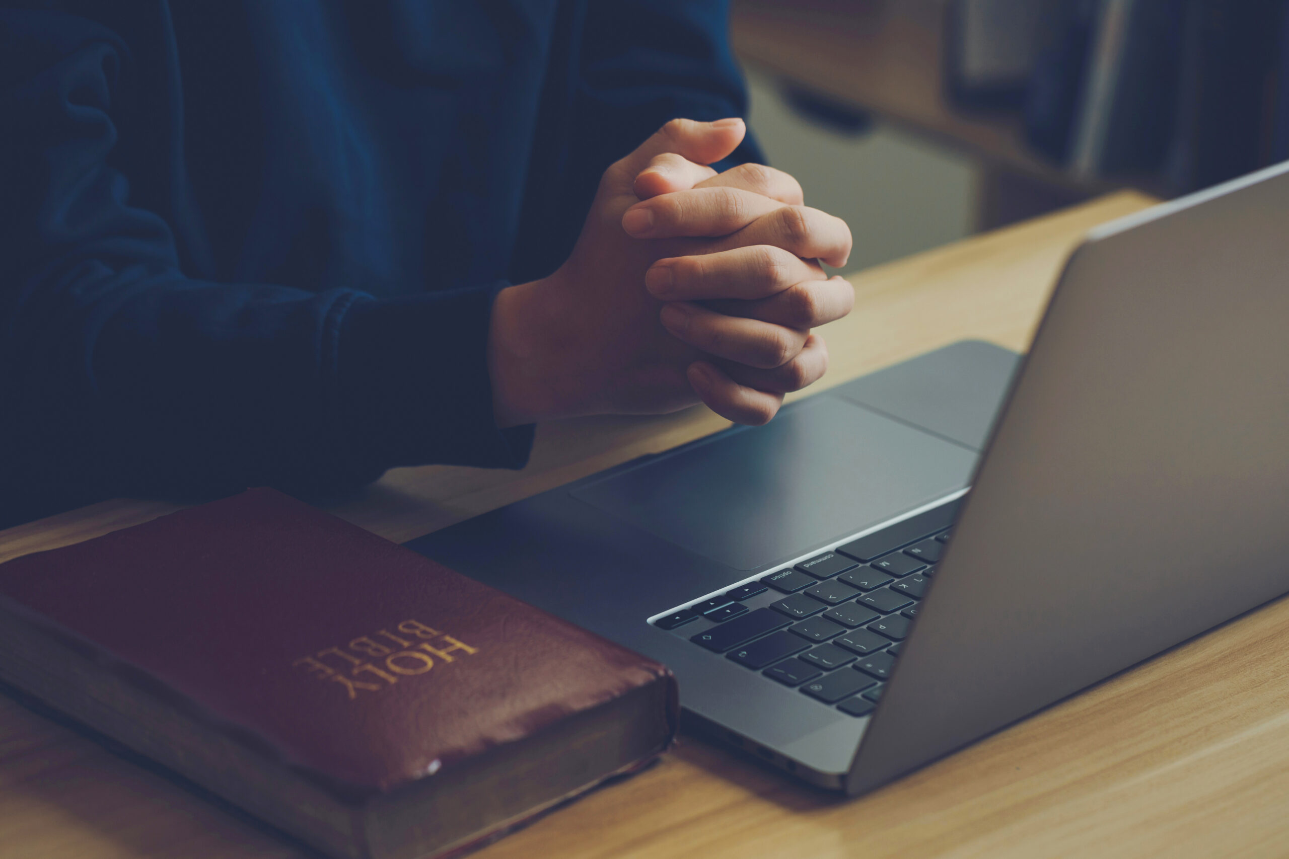 Hands clasped in prayer beside a laptop and Bible on a wooden desk